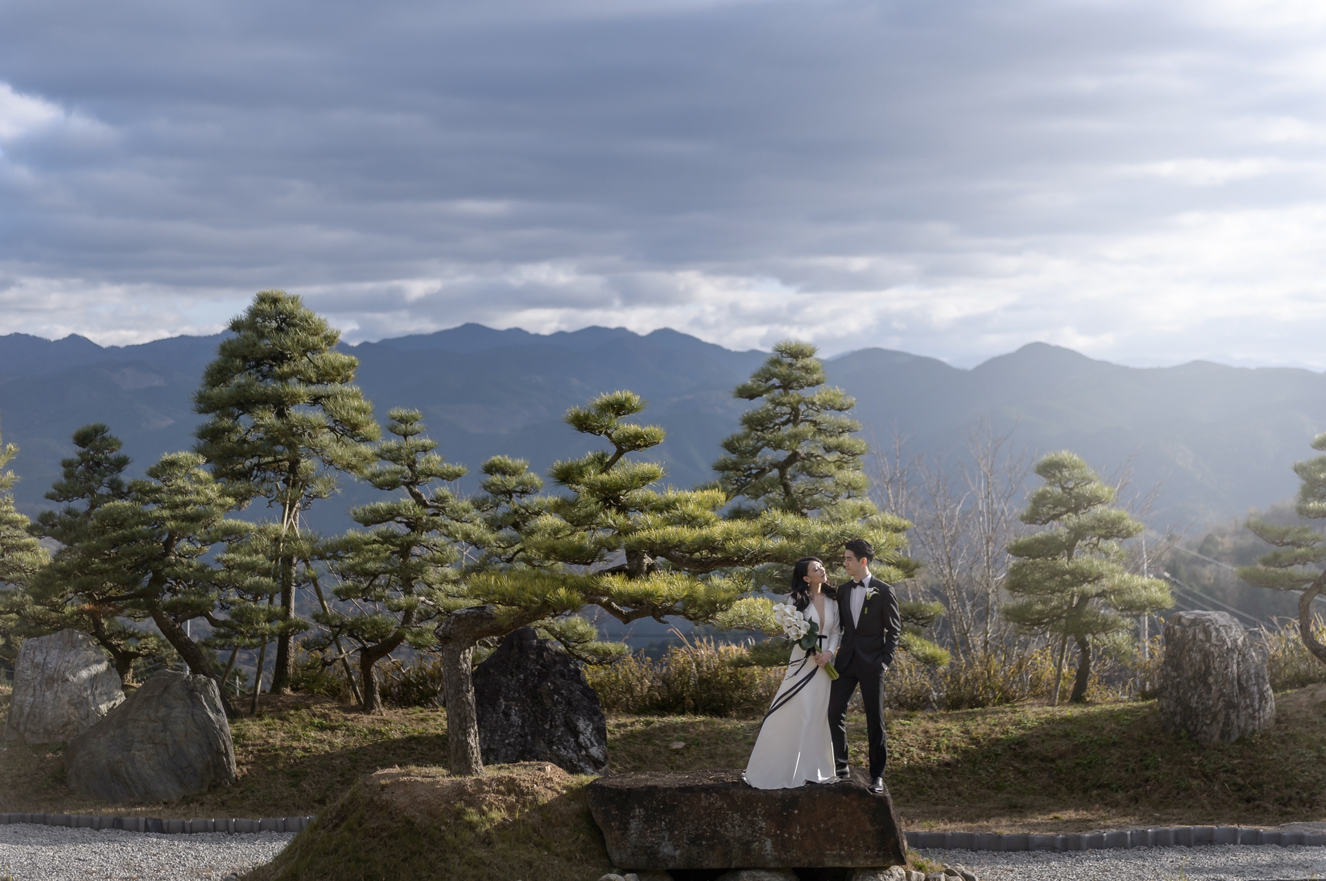 The couple stand on a stone among pruned pines, the mountains of Nara behind them, on the day of their helicopter elopement in Japan.
