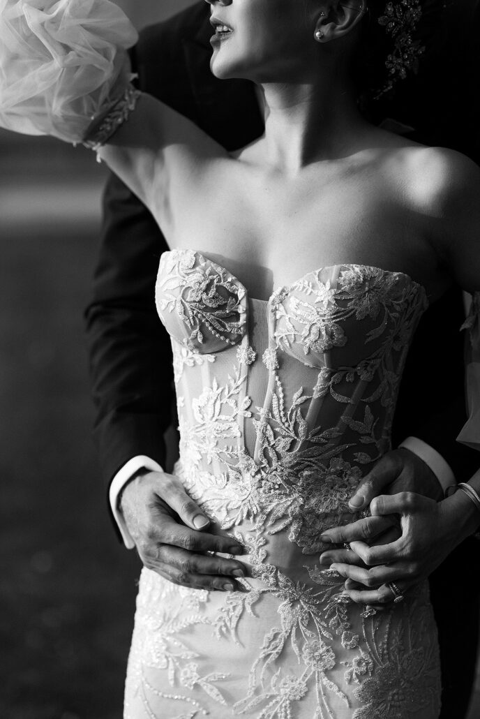 In black and white, the groom's hands rest at the bride's waist against the lace of her strapless gown.