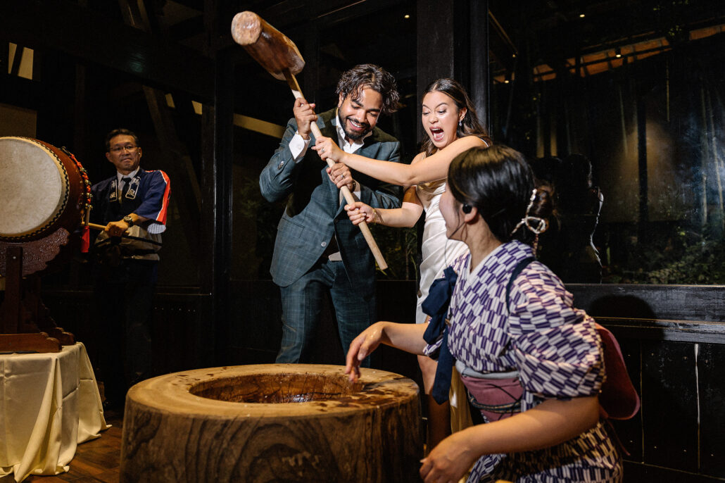 The groom swings a wooden mallet into a mochi mortar as the bride cheers and a woman in kimono waits to turn the rice.