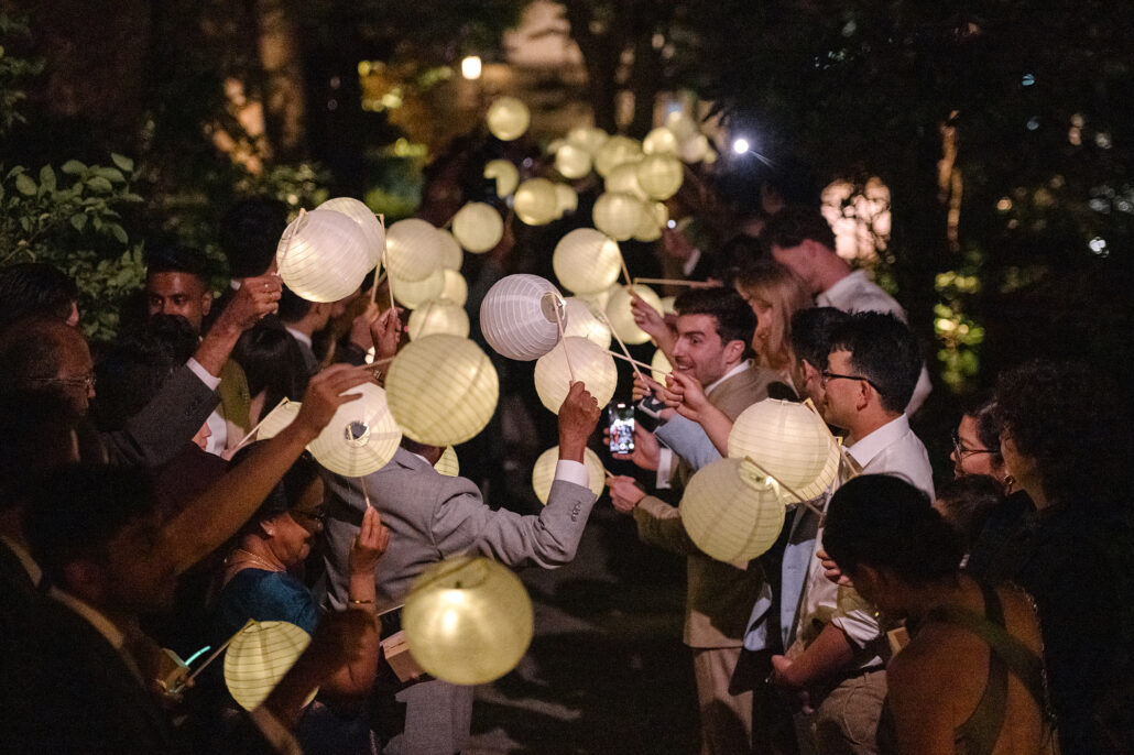 Guests lift glowing paper lanterns into the night around the bride and groom in a garden.