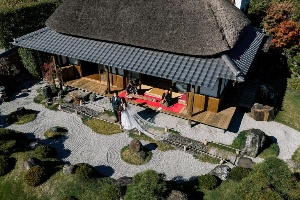 Seen from above, the bride and groom stand in a raked gravel garden in front of a thatched-roof Japanese house with red felt on its veranda.