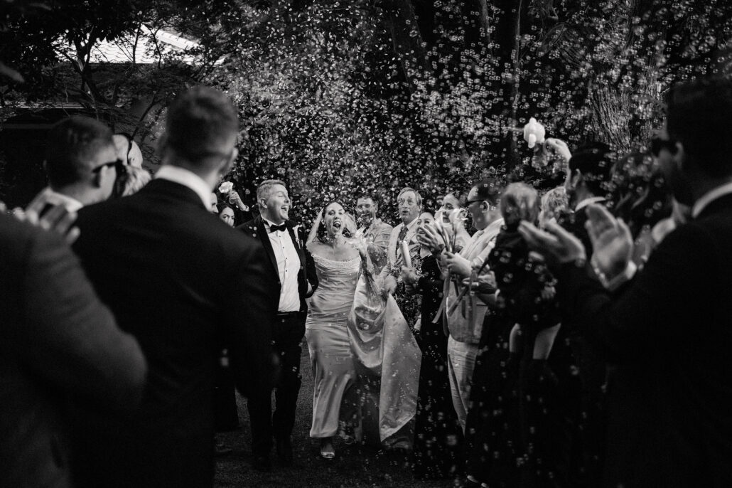 In black and white, the bride and groom walk out through their cheering, clapping guests in a garden.