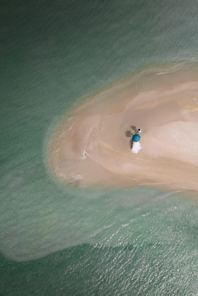 Seen from above, the bride and groom stand on a curving sandbar in a pale green sea.