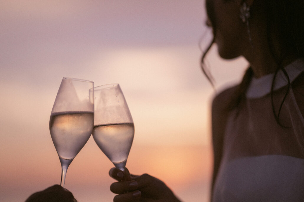 Two glasses of champagne meet against a sunset sky, the bride soft in the background in a halter-neck dress.