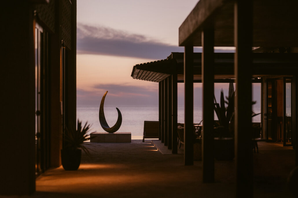 A crescent sculpture stands at the end of a stone terrace above the sea at dusk, between dark timber pavilions.