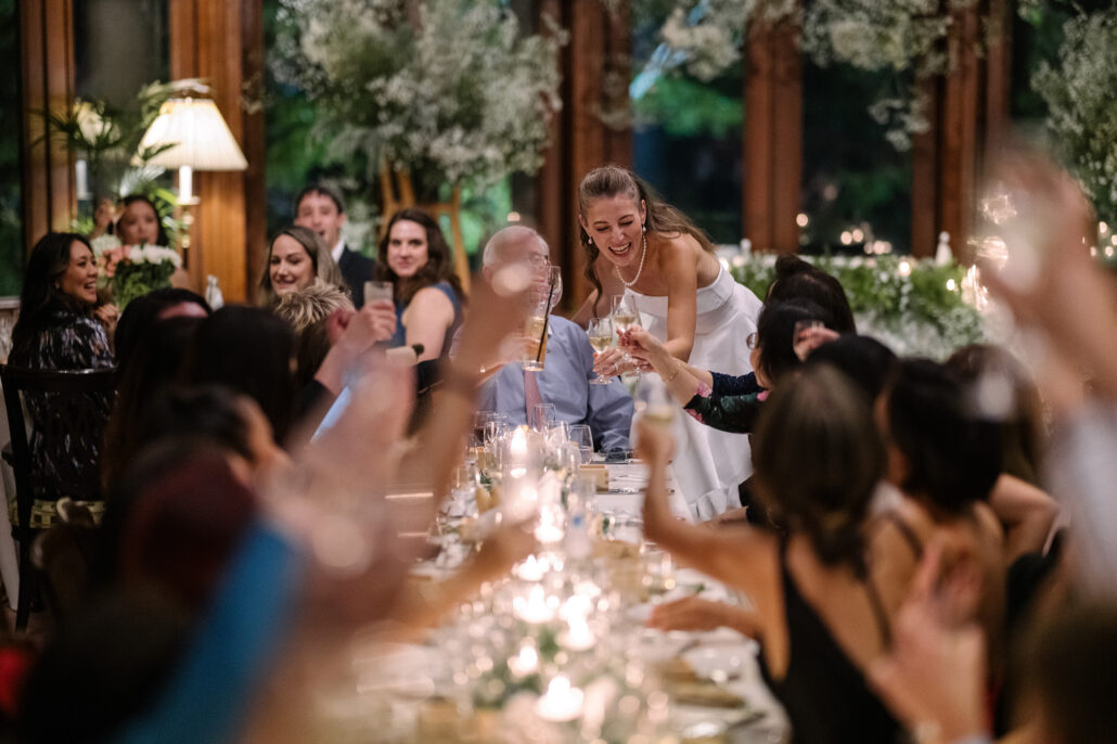 Guests raise their glasses across a candlelit table as the bride laughs, greenery hanging overhead.