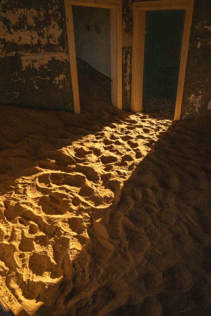 Interior of a Kolmanskop house with teal paint, broken windows, and layered dust.