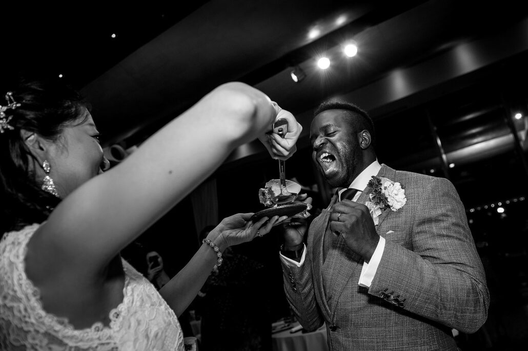 Bride playfully feeds groom cake as he laughs, candid documentary reception moment in black and white.