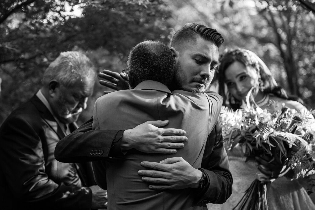 Groom embraces the bride's father in soft afternoon light, emotion etched across both faces, modern documentary style.