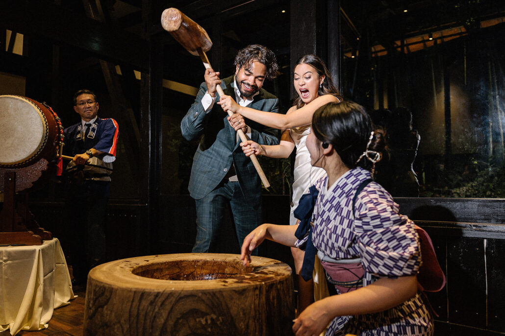Mochi-pounding at a wedding reception with taiko drum, couple laughing mid-swing, Japanese tradition meets party.