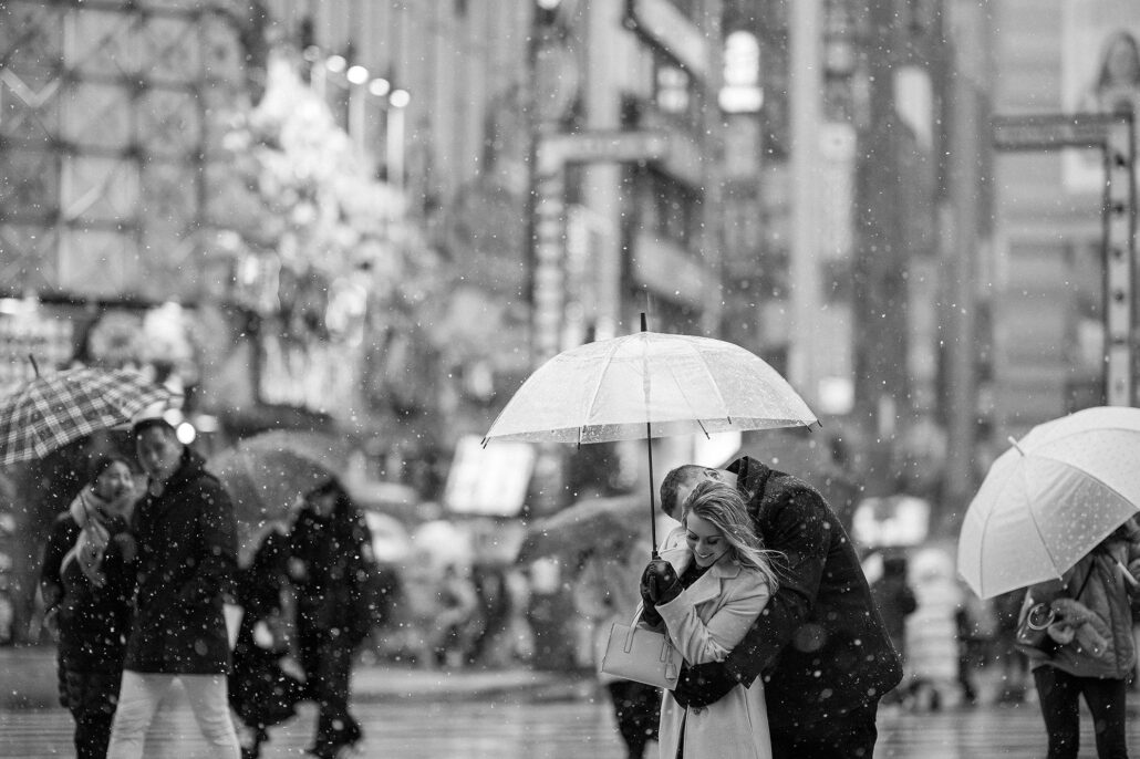 Tokyo street engagement in snowfall, couple under clear umbrella, cozy embrace, documentary style romance.