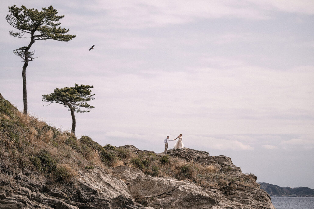Newlyweds on windswept Japanese coastline, adventure portrait beneath pines, location-led storytelling.