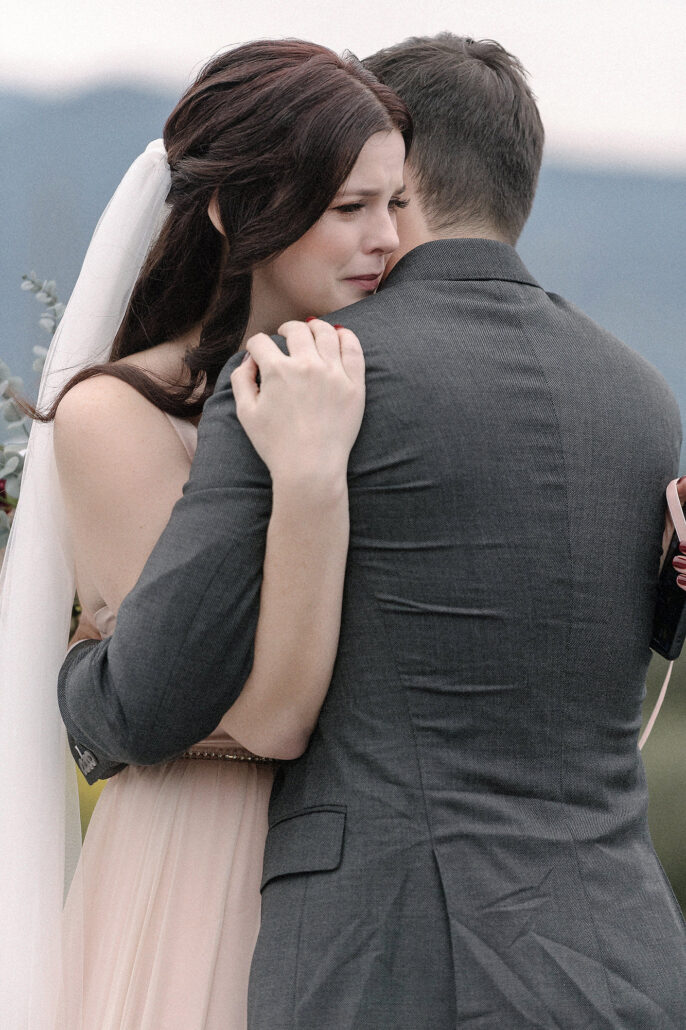 Bride clings to groom with happy tears against soft mountain horizon, intimate, emotive elopement embrace.