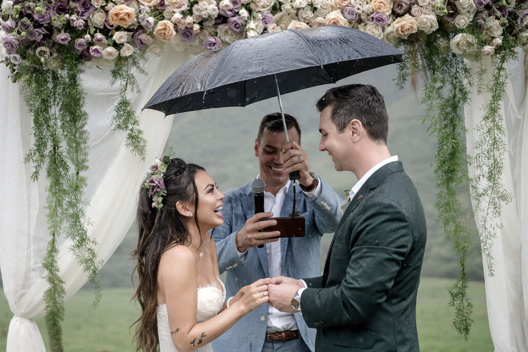 Rainy outdoor wedding ceremony with couple laughing under umbrella, enhanced using AI editing software for photographers to keep detail and warmth in wet weather.