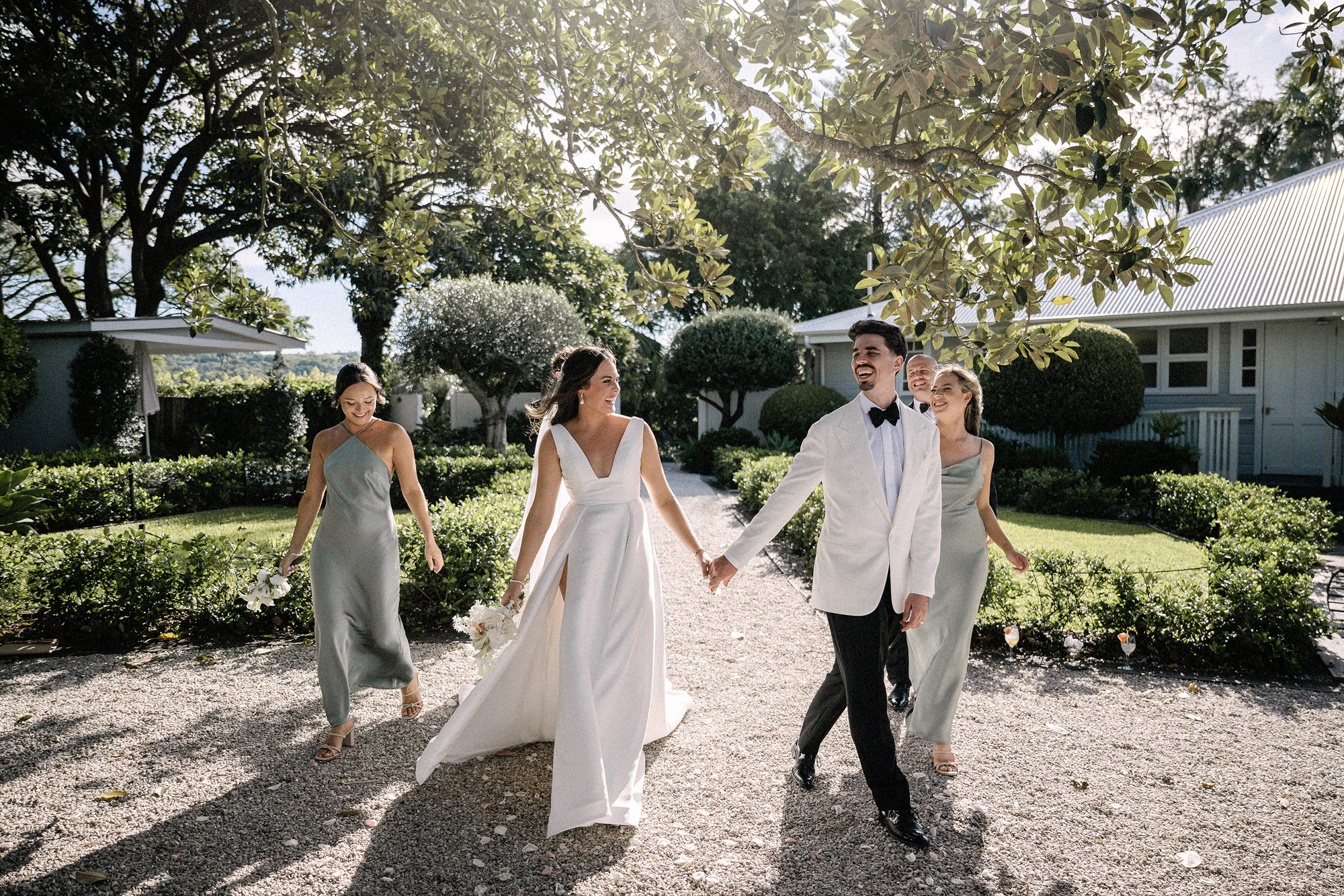 The bride and groom walk hand in hand across the gravel with their bridesmaids beneath the trees at the Fig Tree Restaurant in Byron Bay.