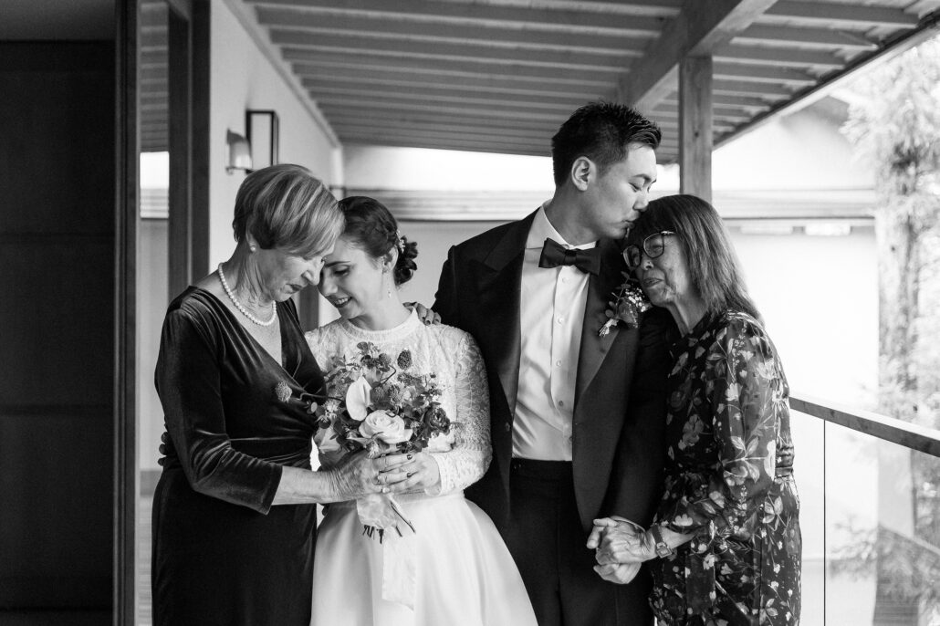 In black and white, the bride and groom stand close with both of their mothers on a covered terrace at the ryokan.