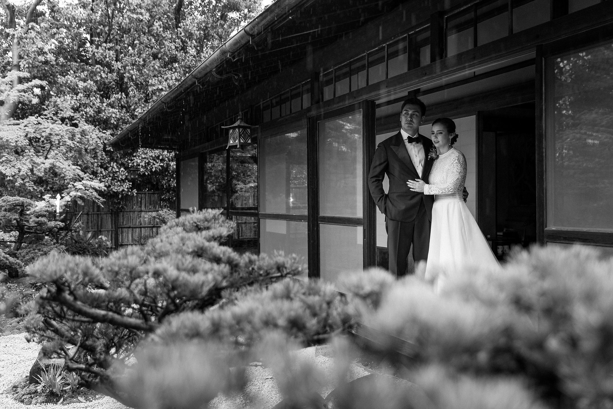 In black and white, the bride and groom stand on the veranda of Jinya Ryokan as rain falls over the pine garden on their wedding day.