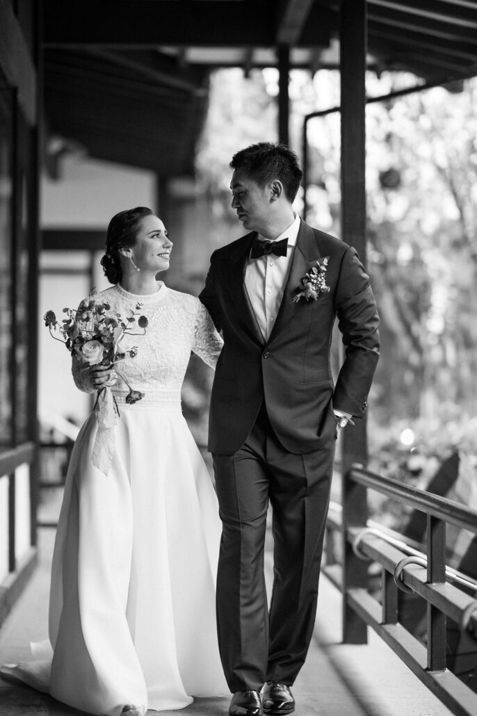 In black and white, the bride and groom walk arm in arm along a covered wooden walkway at their ryokan wedding in Japan.