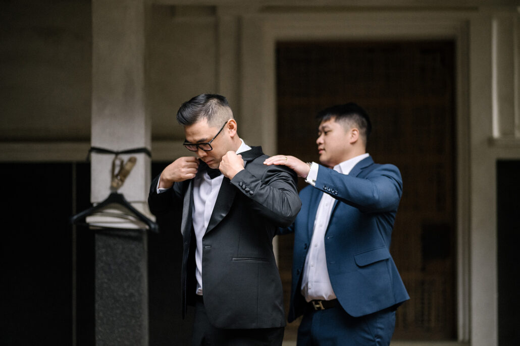 A friend in a blue suit helps the groom into his black tuxedo jacket in the doorway of the old villa in Matsumoto.