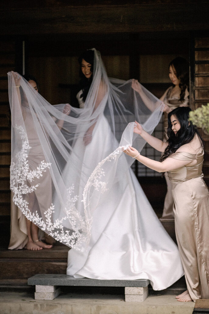 Two bridesmaids in champagne satin lift the bride's long lace veil in a wooden doorway at the villa in Matsumoto.