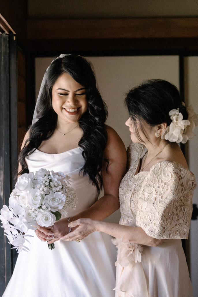 The bride holds white roses beside a woman in a lace dress with a flower in her hair, getting ready in Matsumoto.