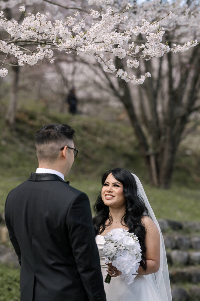 The bride smiles at the groom holding white roses, the cherry trees and stone terraces of the farm behind them in Nagano.