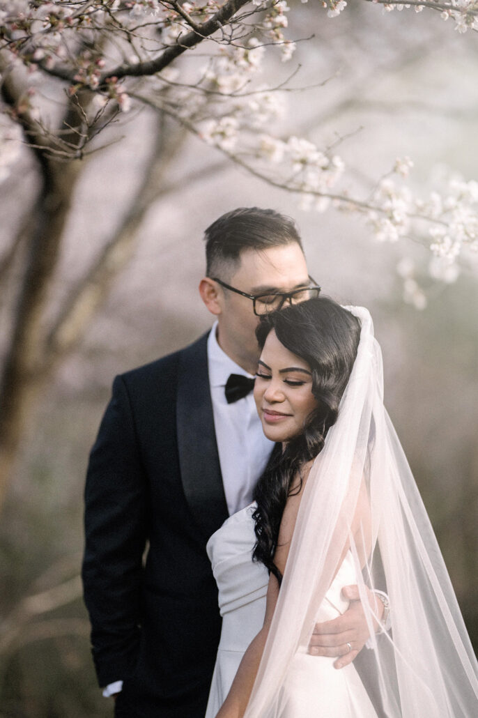 The groom holds the bride close among soft pink cherry blossom, her veil falling behind her, at a Nagano wedding.