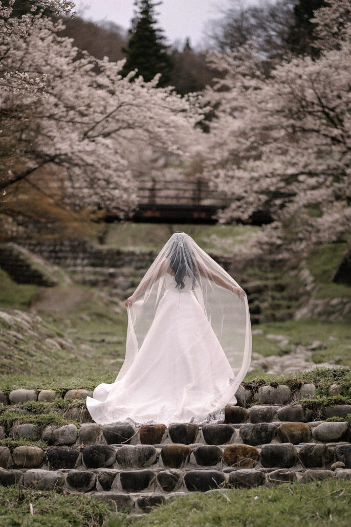 The bride stands on old stone steps with her veil spread wide, a valley of cherry trees ahead, at a Nagano wedding.