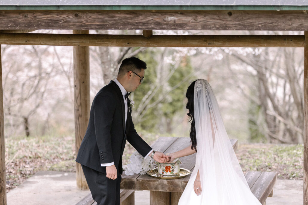 The groom takes the bride's hand across a small tray on a wooden table beneath a pavilion among cherry trees in Nagano.