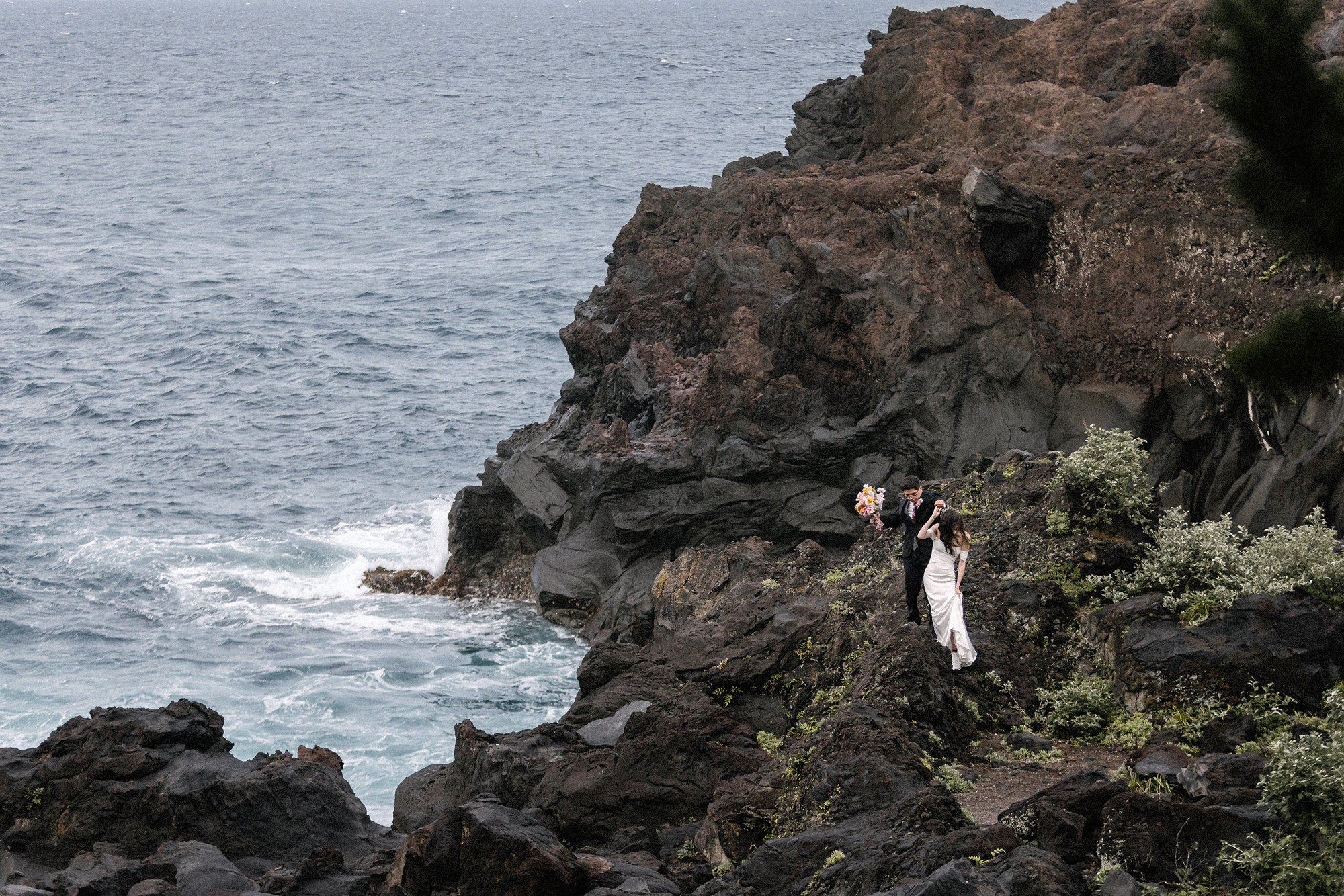 The couple stand on a ledge of dark volcanic rock above the sea, the bride's bouquet raised, at their Izu Peninsula elopement in Japan.