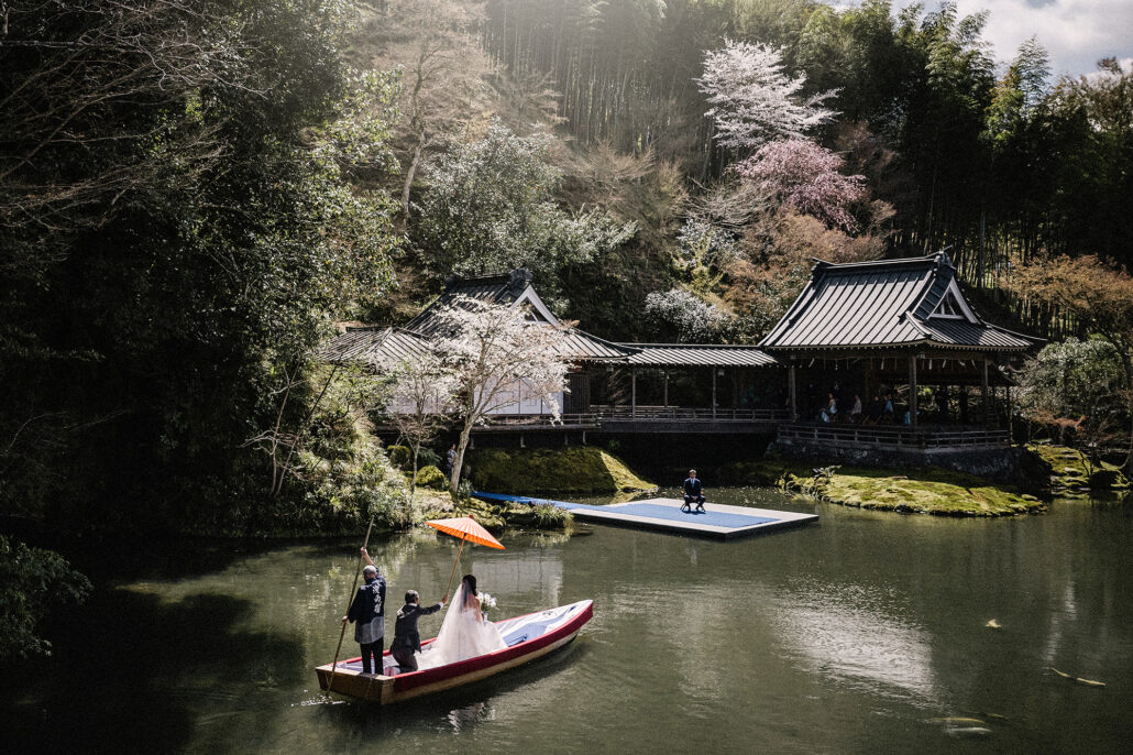 At Asaba, a boatman poles the bride and groom across the pond beneath a red umbrella, toward the floating noh stage and spring blossom on the hill.