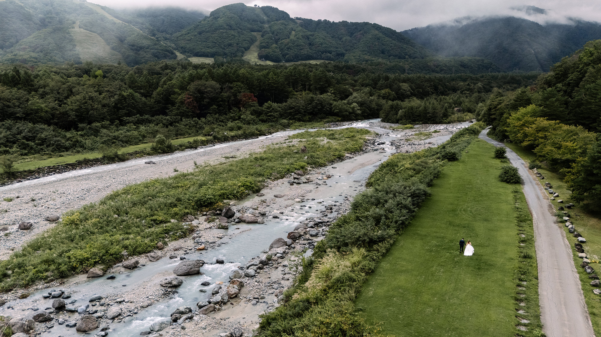 A drone view of the bride and groom on a long green meadow beside a stony river, below misty ski slopes in Hakuba, Japan.