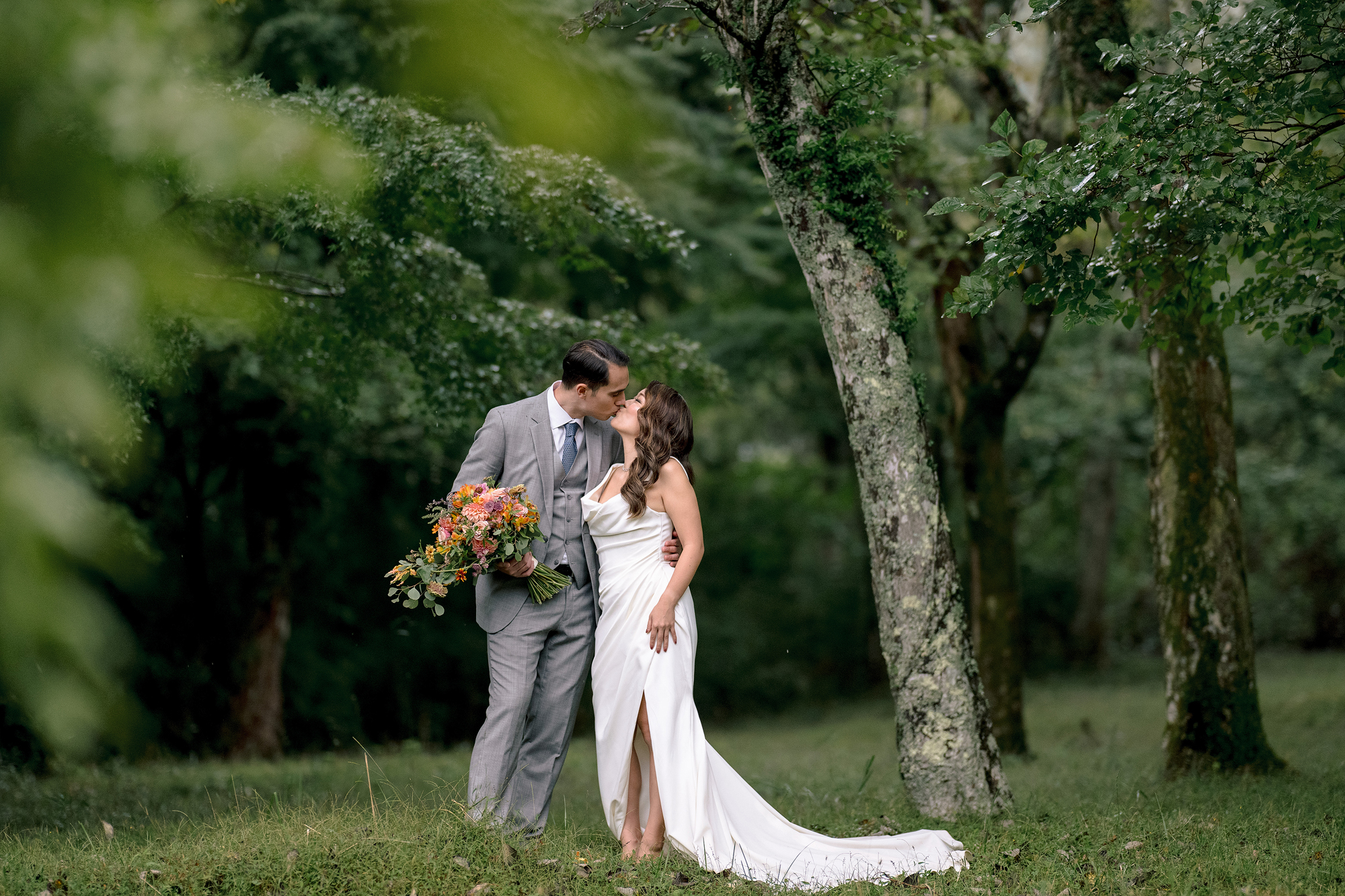 The couple kiss among mossy trees in the Hakone forest, the groom holding her bouquet of autumn flowers on their wedding day.