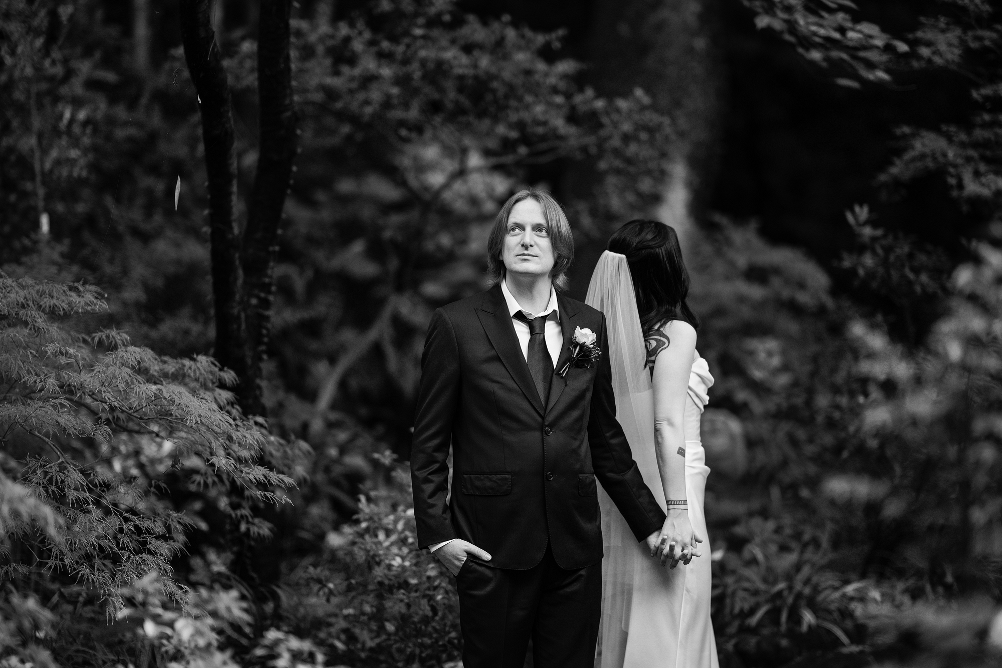 In black and white, the groom looks ahead as he holds the bride's hand in the green garden of Jinya Ryokan on their wedding day.