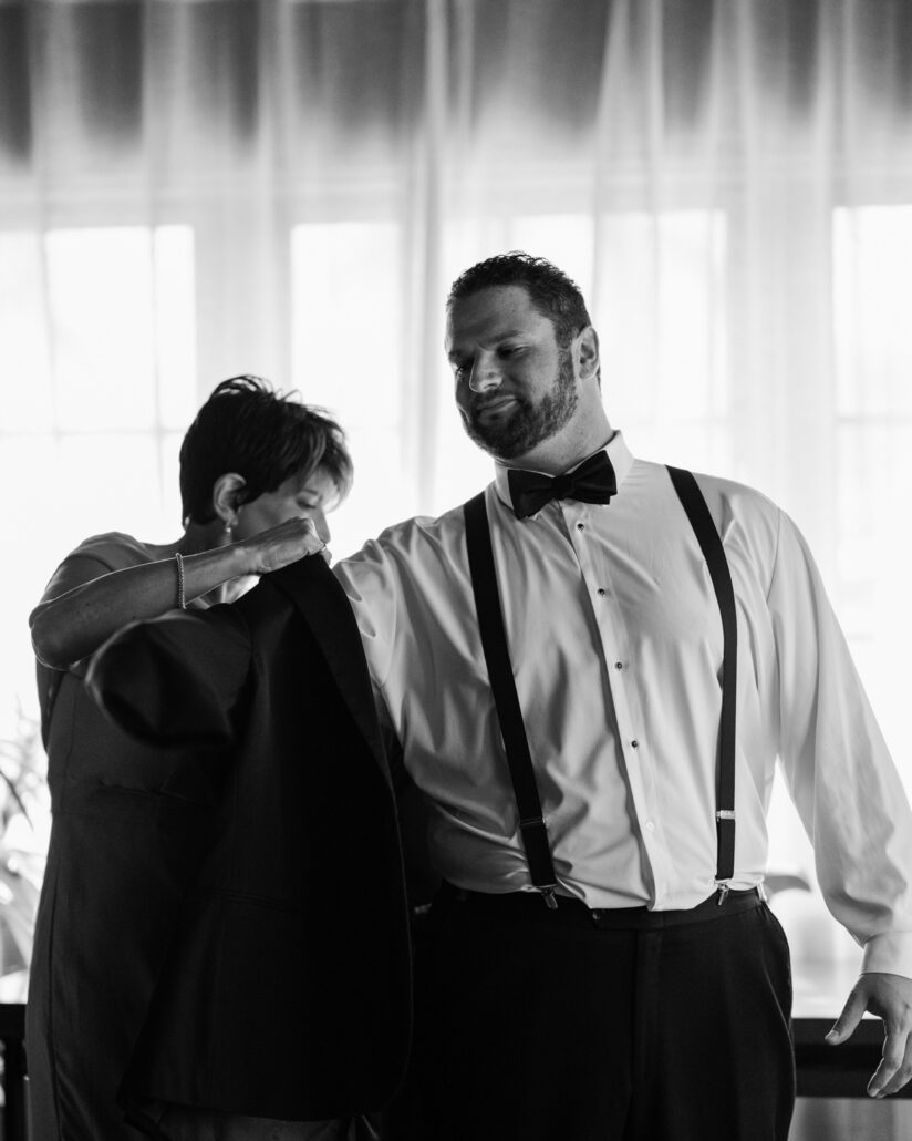 In black and white, a woman helps the groom into his jacket, her head bowed close to it, as he smiles down at her before his Florida Keys wedding.