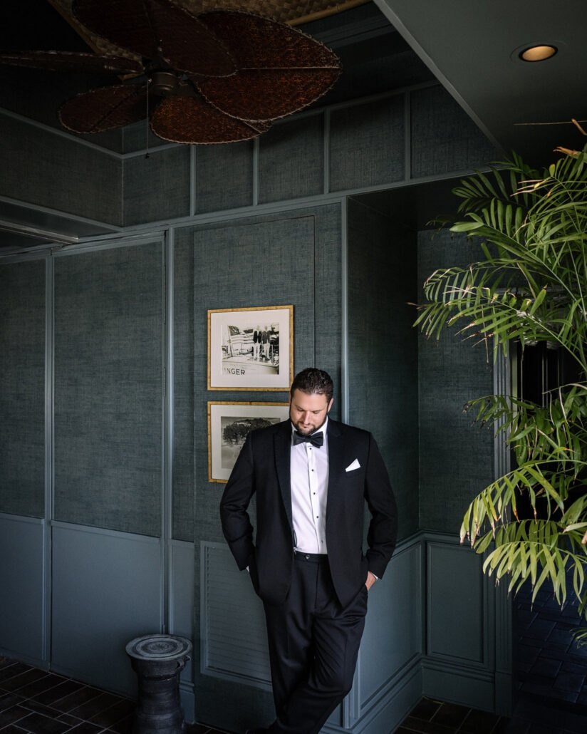 The groom leans against a green panelled wall beside a leafy palm, a rattan ceiling fan overhead, ready for his Little Palm Island wedding.