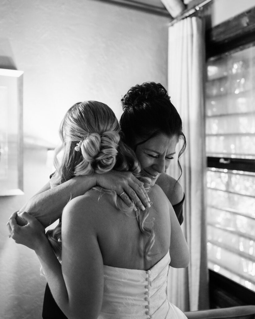 In black and white, a woman hugs the bride by the window with her eyes closed, the buttons down the back of the gown in view, before a Florida Keys wedding.