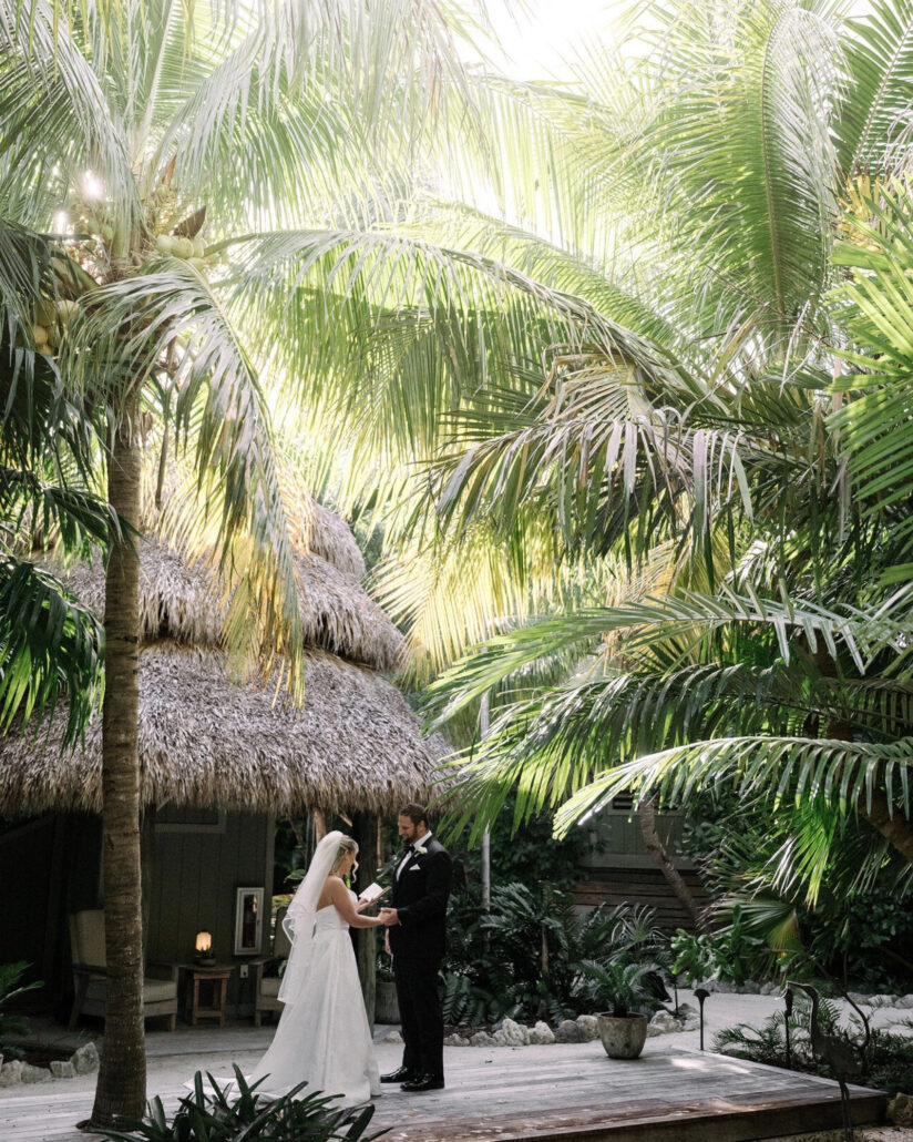 Tall coconut palms and a thatched roof frame the groom as he reads his vows to the bride on a wooden deck, a private island wedding in the Florida Keys.