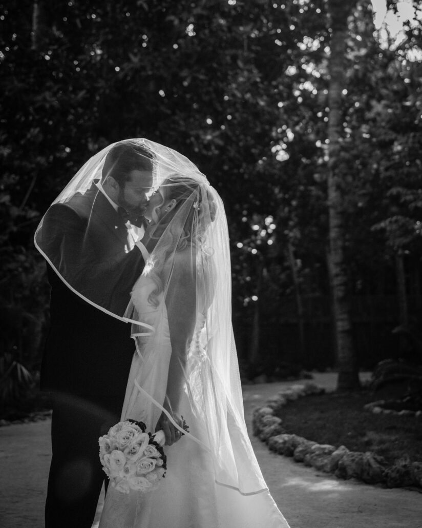 In black and white, the groom kisses the bride under her veil on a winding garden path at their Little Palm Island wedding.