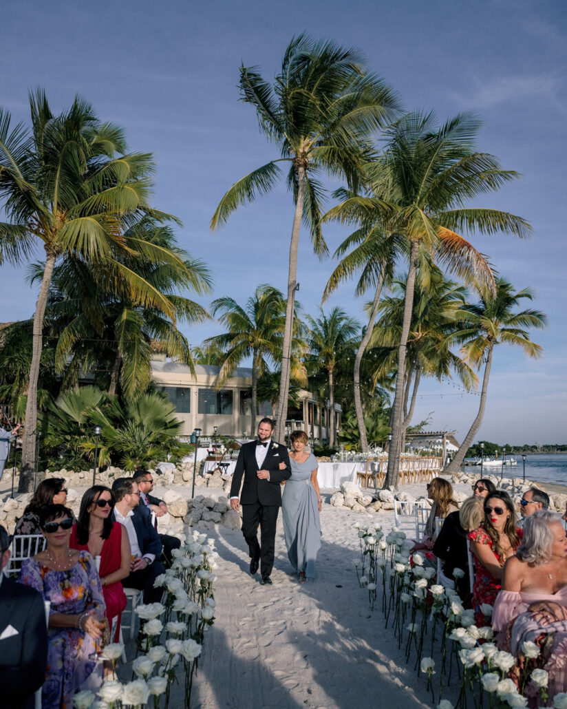 The groom walks a woman in a pale blue gown down a sandy aisle lined with white roses, tall palms overhead at a Florida beach wedding.