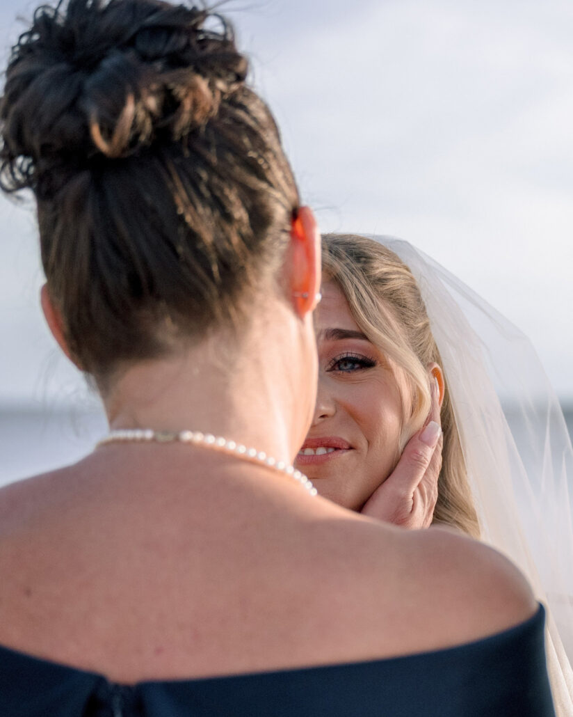 Seen over her shoulder, a woman in a pearl necklace cups the bride's cheek in her hand at the beach wedding ceremony on Little Palm Island.