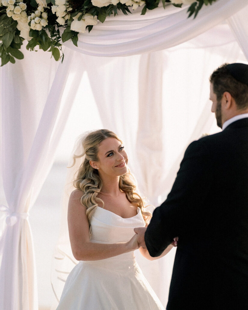 The bride smiles up at the groom in his kippah as they hold hands beneath a white draped arch crowned with roses at their Florida Keys beach wedding.