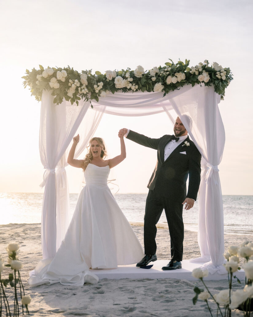 The bride and groom raise their joined hands and cheer under the floral arch, the sea bright behind them at their beach wedding in the Florida Keys.