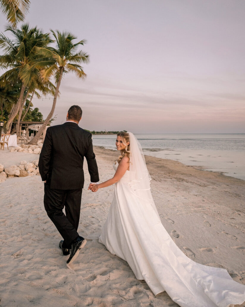 The groom leads the bride along the sand past leaning palms at dusk, her long train trailing as she looks back over her shoulder on a Florida Keys beach.