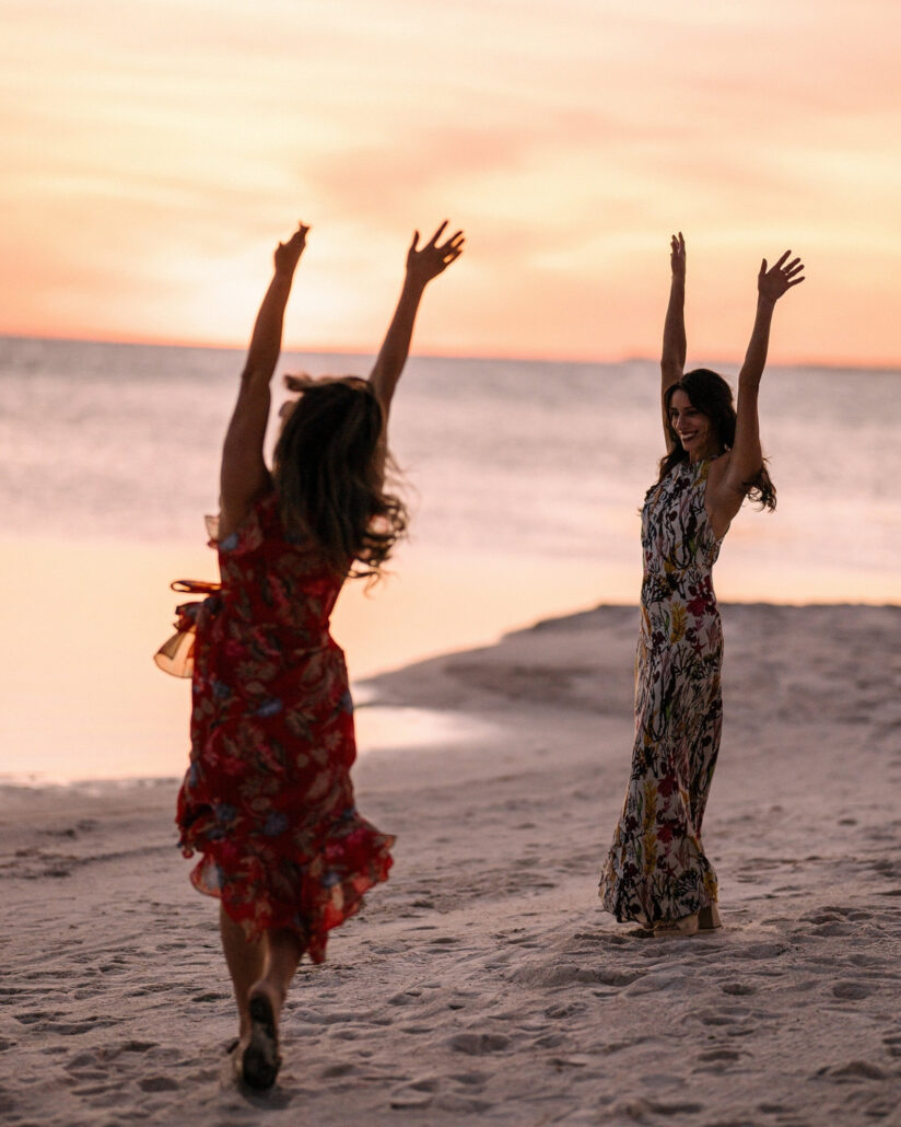 Two guests throw their arms up on the beach at sunset, one caught mid stride in a red floral dress, at a destination wedding in the Florida Keys.