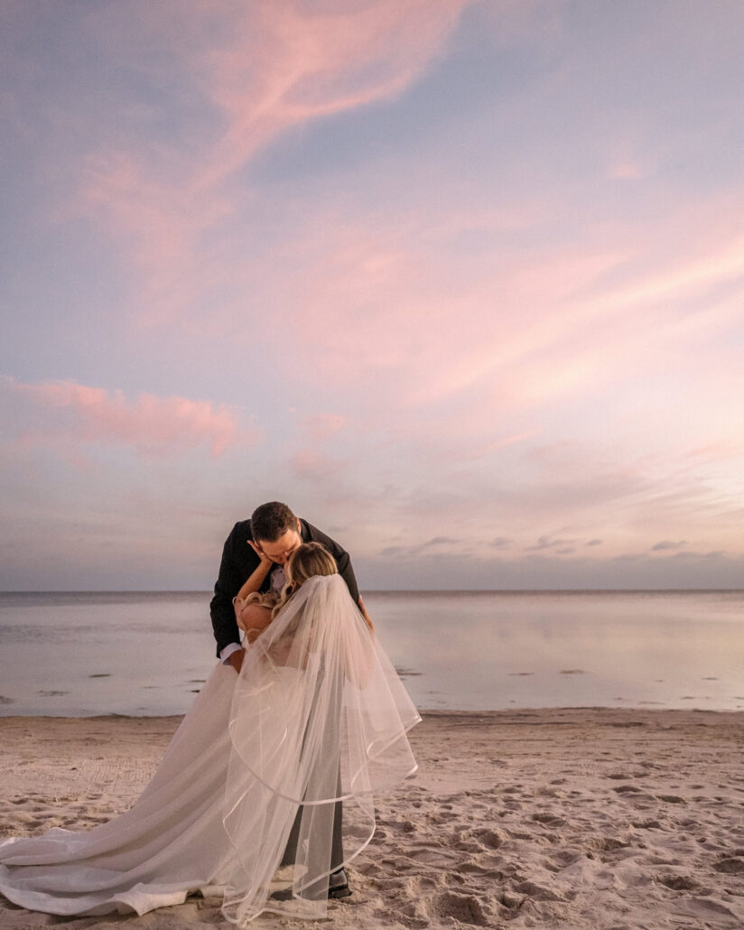 Under a pink streaked sky, the groom bends to kiss the bride on the beach as her veil and train spill across the sand at their Little Palm Island wedding.