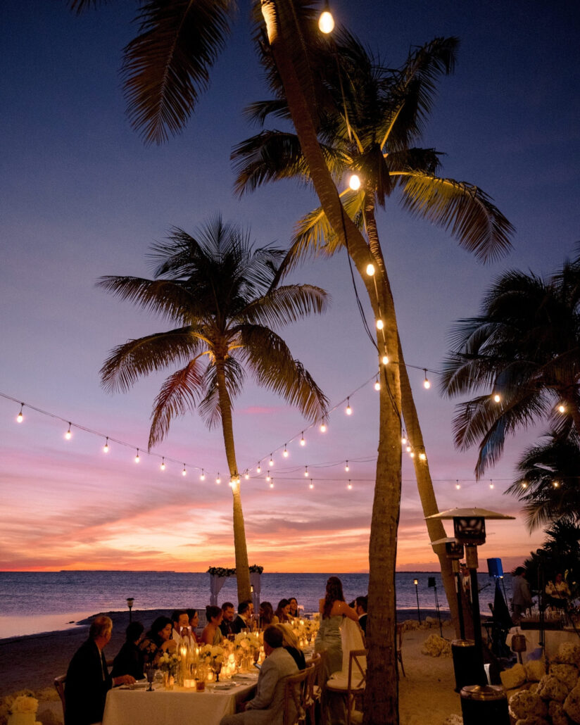 String lights run between tall palms over candlelit dinner tables on the sand as the sunset fades pink and violet over the water at Little Palm Island.