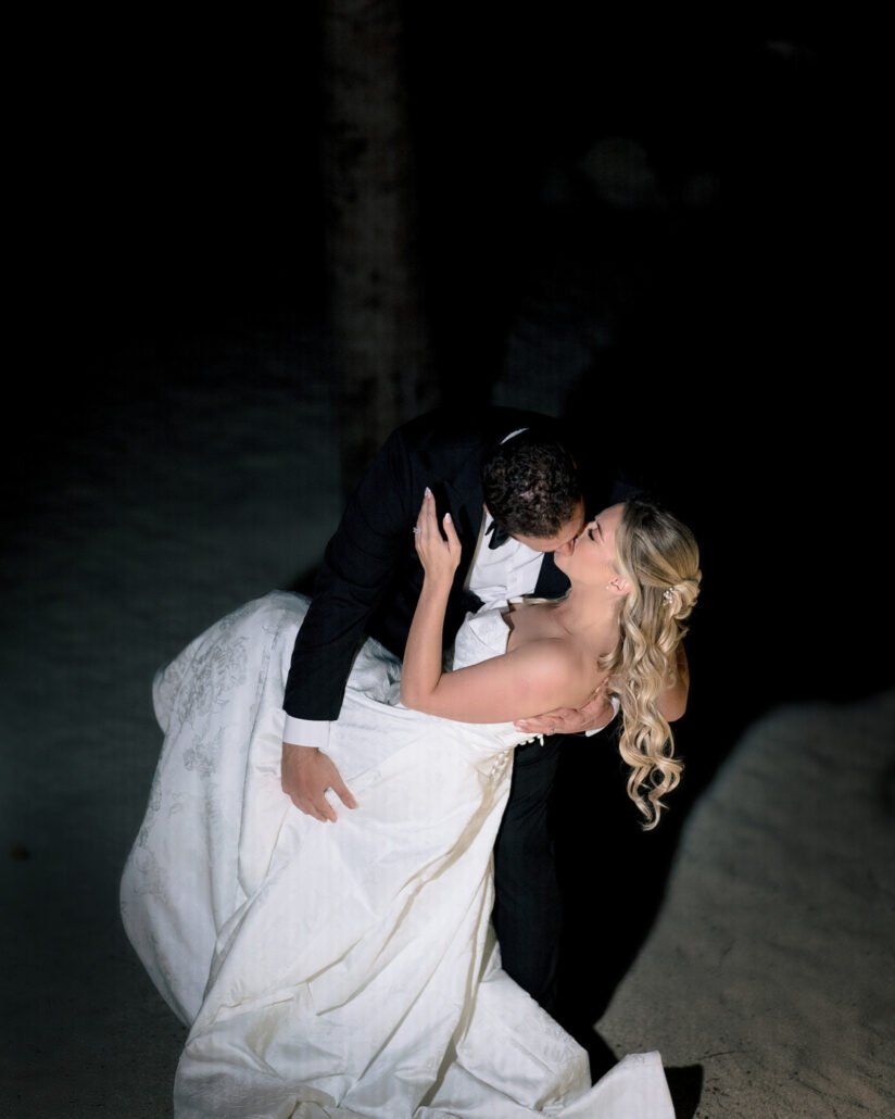At night on the beach, the groom dips the bride into a kiss, her gown pooling on the sand and her hand at his cheek, at their Florida Keys wedding.