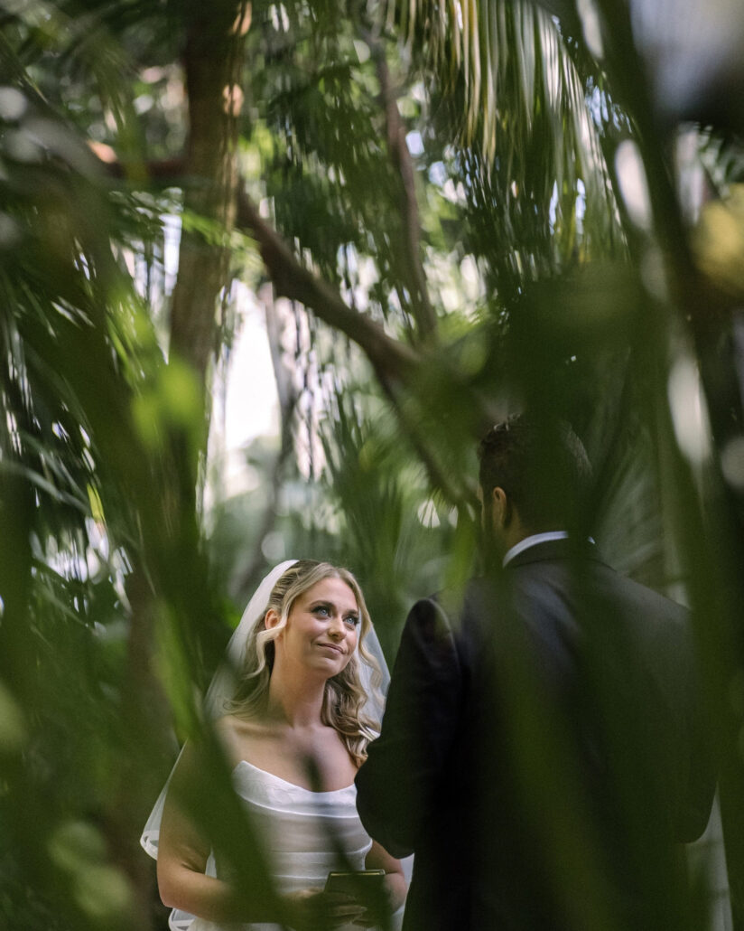 The bride looks up at the groom through a screen of palm leaves as they share private vows in the gardens of Little Palm Island.