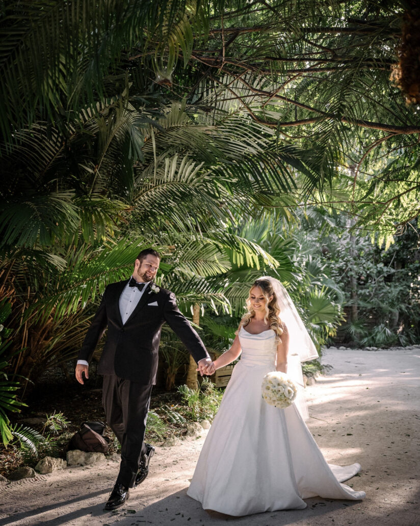 The groom smiles as he leads the bride by the hand along a sandy path under palm fronds, sunlight falling across her gown on Little Palm Island.